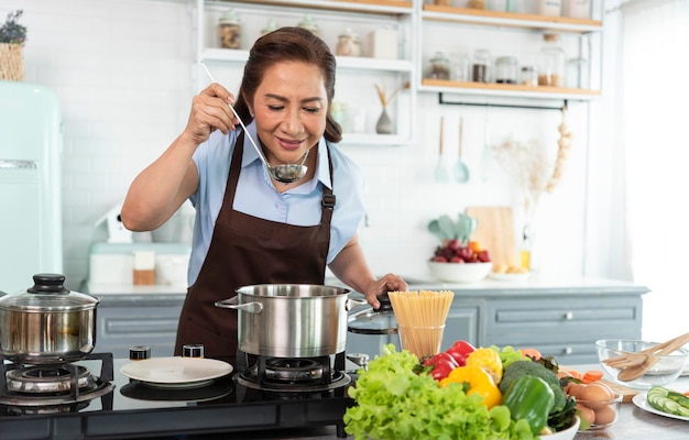 Happy smiling asian older woman in apron is tasting and smell food while cooking in kitchen