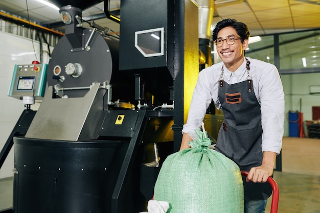 Proprietario asiatico sorridente felice della torrefazione che spinge il carrello con il sacco di caffè tostato