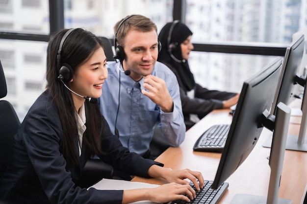 Call center ed operatore asiatici sorridenti felici della donna con le cuffie avricolari d'uso del collega che lavorano al computer e che parlano con il cliente con la sua mente di servizio