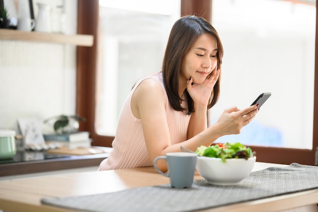 Donna asiatica sorridente felice che utilizza il cellulare dello smartphone a casa in cucina con un'insalatiera sul tavolo