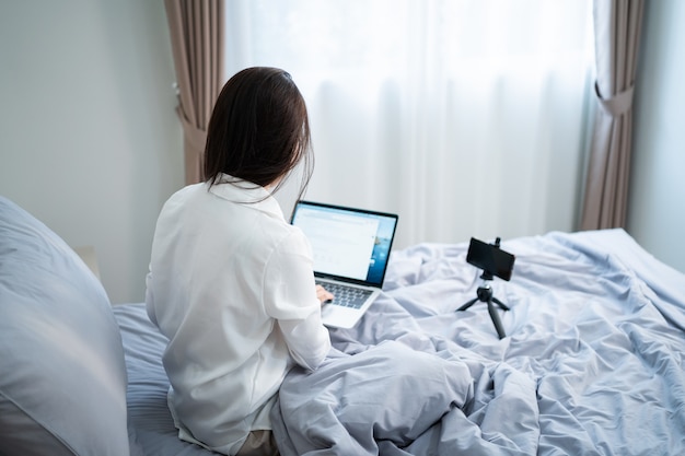 Счастливая улыбающаяся азиатская молодая женщина с портативным компьютером и мобильным телефоном с видеозвонком в спальне