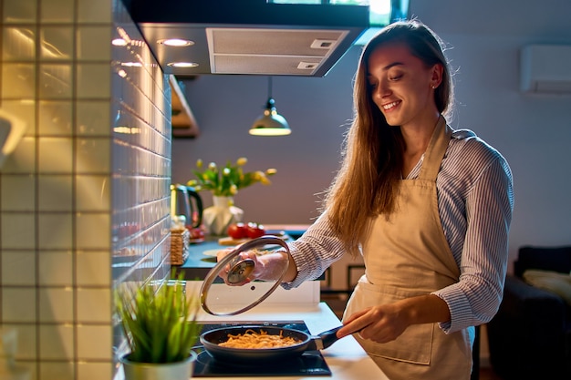 Счастливая улыбающаяся привлекательная молодая кулинария женщина-домохозяйка готовит еду на сковороде на плите для вечернего ужина на современной кухне в стиле лофт