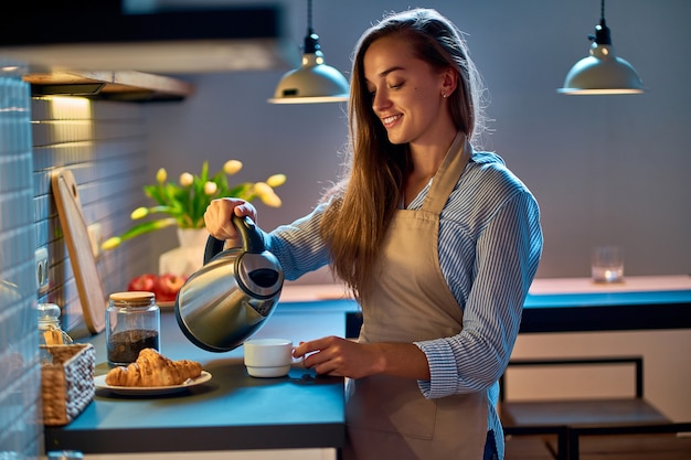 Счастливая улыбающаяся привлекательная молодая женщина-домохозяйка наливает и заваривает чай, используя электрический чайник для вечернего перерыва на кофе на современной кухне в стиле лофт