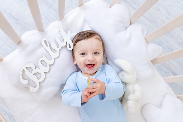 Happy smiling baby boy in the crib with the inscription baby in a blue bodysuit, cute joyful little baby in the bedroom