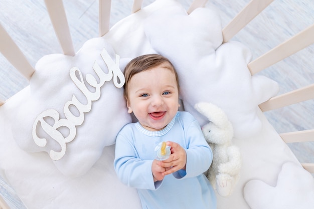 Happy smiling baby boy in the crib with the inscription baby in a blue bodysuit, cute joyful little baby in the bedroom