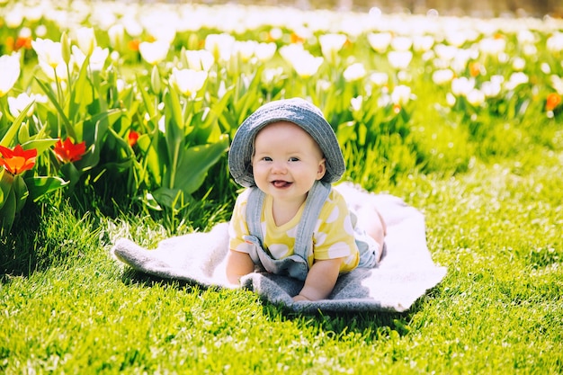 Happy smiling baby girl lying on blanket in green grass of tulips field Child playing outdoors in spring park Image of Mothers Day Easter Family on nature in Arboretum Slovenia Europe