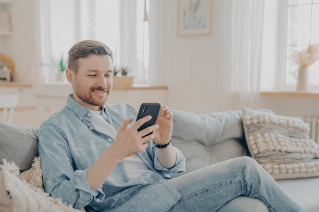 Uomo barbuto sorridente felice che utilizza il dispositivo smartphone mentre è seduto sul divano in soggiorno, resta sul divano e legge messaggi sul cellulare mentre si rilassa durante il tempo libero a casa