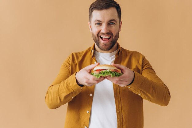 Happy and smiling bearded man in yellow shirt eating burger on beige background