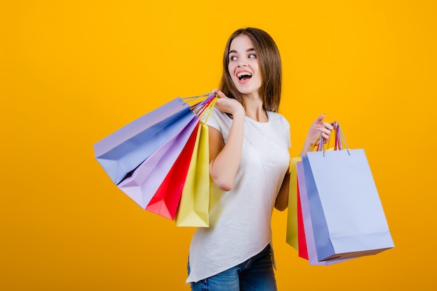 Happy smiling beautiful brunette woman with colorful paper shopping bags wearing jeans and shirt isolated over yellow