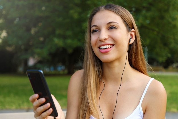 Happy smiling beautiful girl listening music on smart phone enjoying summertime in the park.