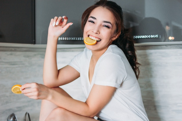 Giovane donna divertente della bella ragazza asiatica sorridente felice dei capelli lunghi con il limone in cucina