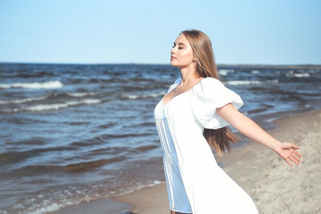 Happy smiling beautiful woman is on the ocean beach in a white summer dress, open arms