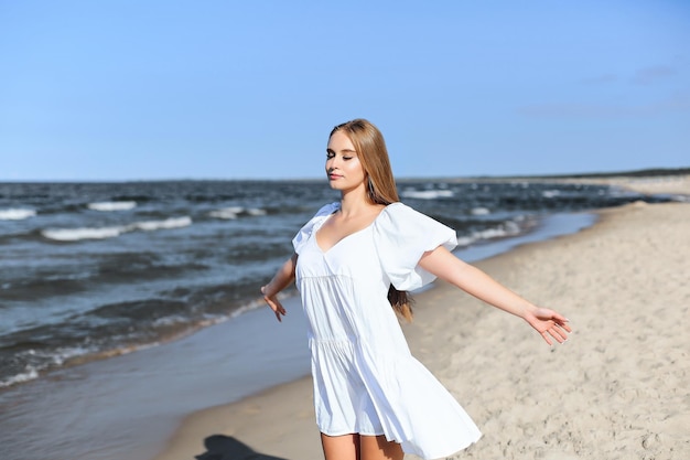 Счастливая улыбающаяся красивая женщина на берегу океана в белом летнем платье с распростертыми объятиями.
