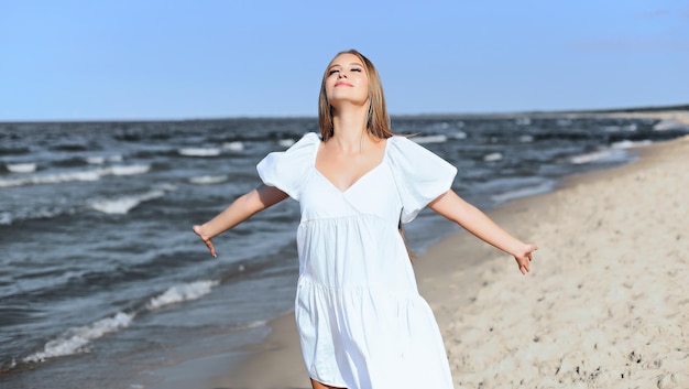 Happy smiling beautiful woman is on the ocean beach in a white summer dress, open arms.