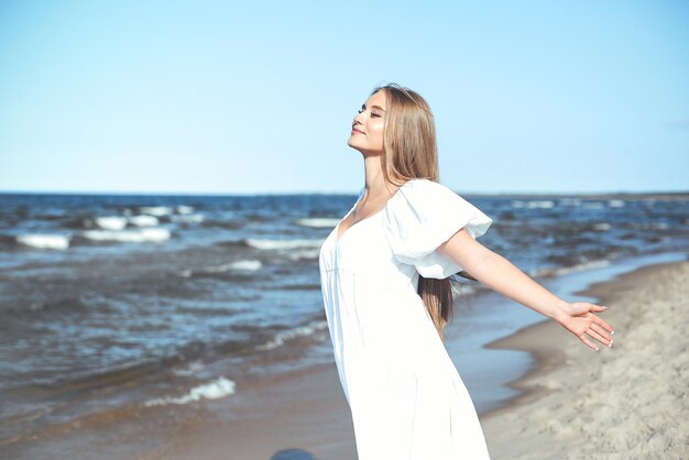 Happy smiling beautiful woman is on the ocean beach in a white summer dress open arms