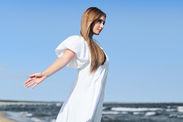 写真 幸せな笑顔の美しい女性は白い夏のドレスを着て海辺にいて腕を開けています