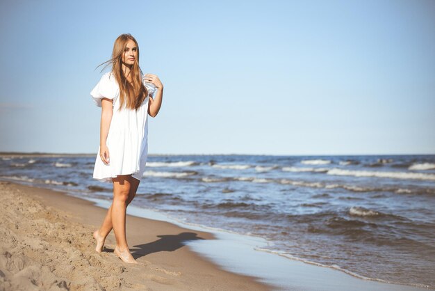 Счастливая улыбающаяся красивая женщина гуляет по океанскому пляжу в белом летнем платье