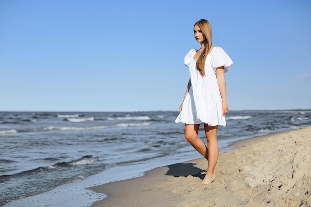 Happy smiling beautiful woman is walking on the ocean beach in a white summer dress