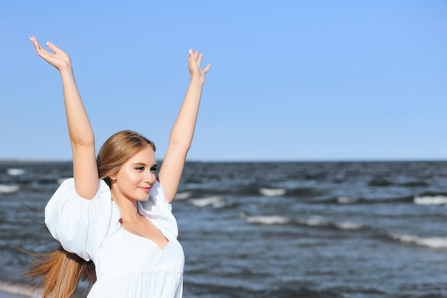 Happy smiling beautiful woman on the ocean beach standing in a white summer dress, raising hands.