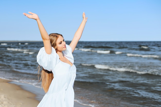 Happy smiling beautiful woman on the ocean beach standing in a white summer dress, raising hands.