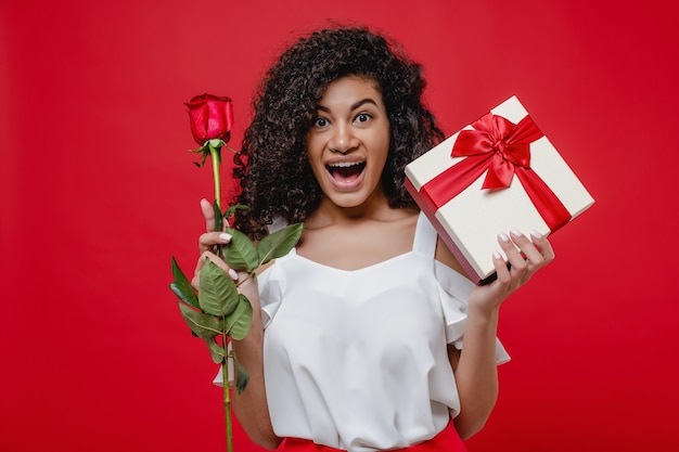 Ragazza nera sorridente felice con il contenitore di regalo e rosa isolato sopra rosso