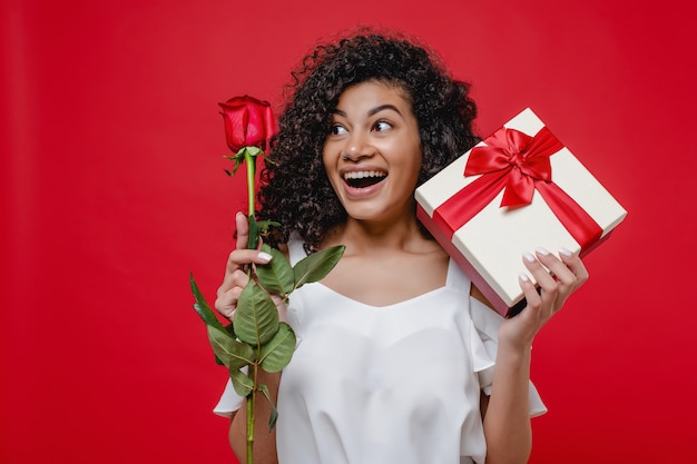 Счастливая усмехаясь черная девушка с розой и подарочная коробка изолированные над красным цветом