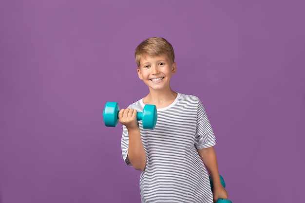 Happy smiling blond boy using fitness equipment holding green dumbbell building arm muscle looking at camera staying on purple background Studio shot