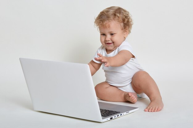 Happy smiling blond curly haired child sitting barefoot on floor with laptop computer and pulls his hands to screen