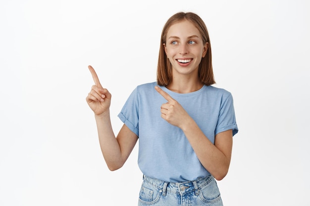 Happy smiling blond woman pointing and looking at upper left corner with cheerful face expression standing in summer tshirt over white background