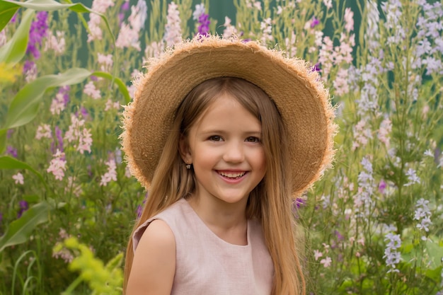 a happy smiling blonde girl in a straw hat and a pink dress is sitting in a flower garden