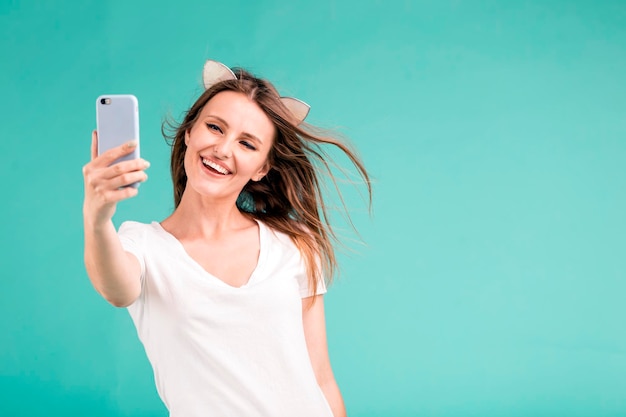 Happy smiling blonde teen girl makes selfie via smartphone on blue background