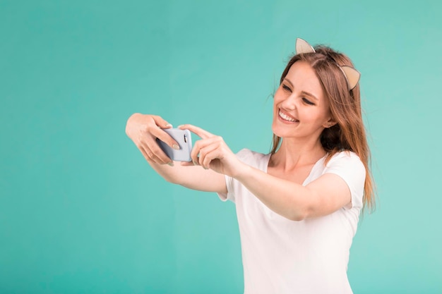 Happy smiling blonde teen girl posing with phone makes selfie via smartphone on blue background