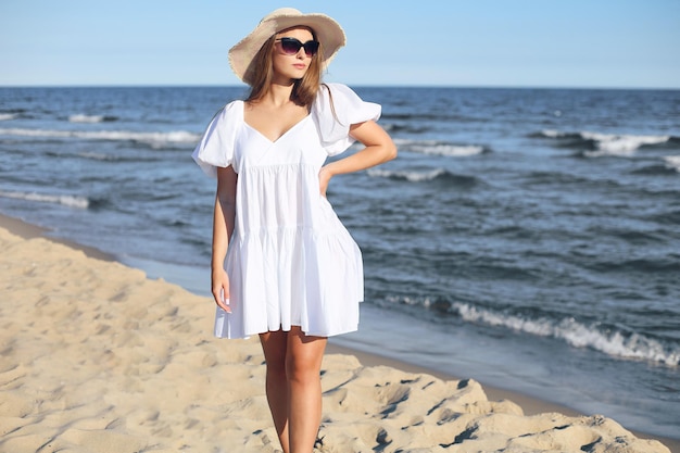 Happy smiling blonde woman is posing on the ocean beach with sunglasses and a hat.