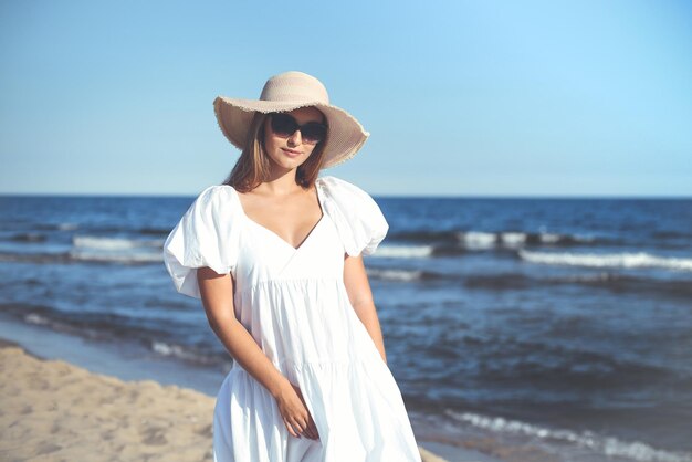 Happy smiling blonde woman is posing on the ocean beach with sunglasses and a hat