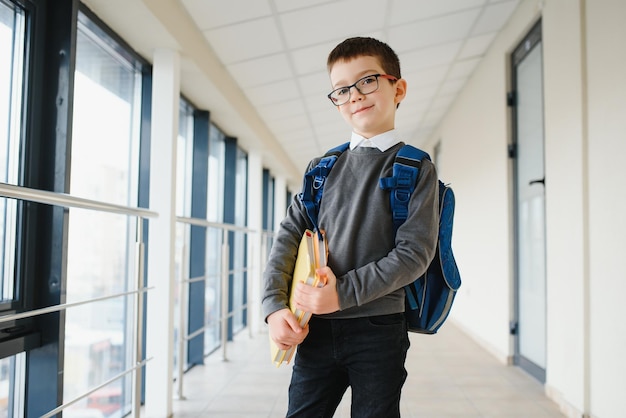 Happy smiling boy in glasses is going to school for the first time Child with school bag and book in his hand Kid indoors of the class room Back to school