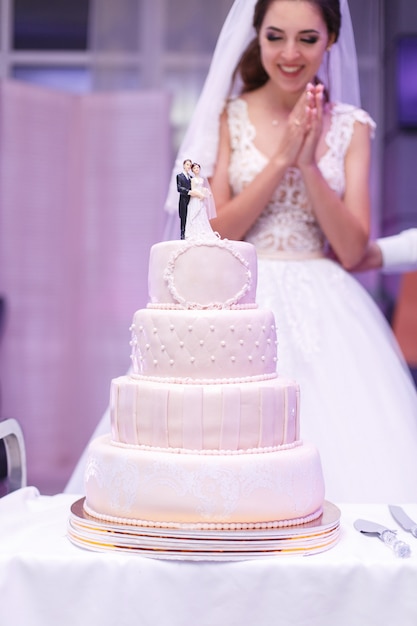 행복 한 미소 신부 레스토랑에서 실내 아름 다운 높은 웨딩 케이크에 의해 서. 하얀 축제 케이크 테이블에 매 스틱 장식.