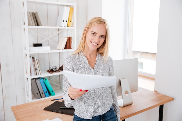 Happy smiling business woman sitting on office desk and giving document to front