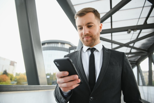 Счастливый улыбающийся бизнесмен в черном костюме и использующий современный смартфон рядом с офисом рано утром успешный работодатель, чтобы заключить сделку, стоя возле офиса небоскреба при ночном свете вспышки