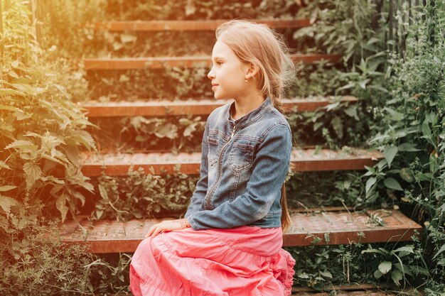 Happy smiling candid eight year old kid girl sits on staircase\
overgrown with grass in nature prepubertal age of children and\
their lives mind psychology and mental health concept flare