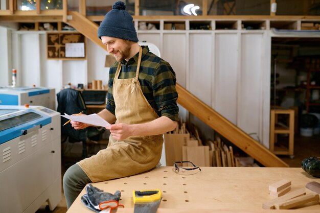 Фото Счастливый улыбающийся плотник, глядя на чертеж, чувствует удовлетворение и успех. профессиональный ремесленник, работающий над проектом в столярной мастерской