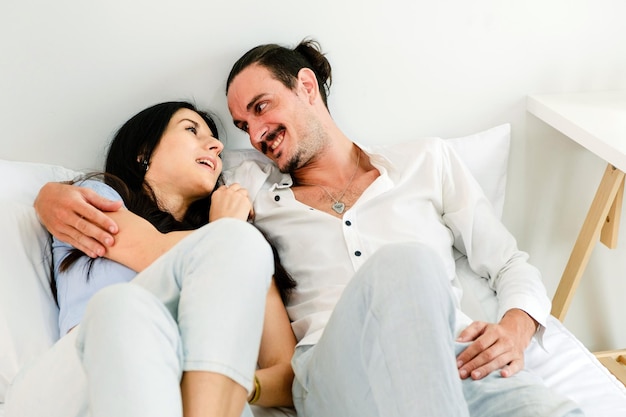 Счастливая улыбающаяся кавказская пара лежит на удобной кровати, расслабляется и смотрит друг другу в глаза.