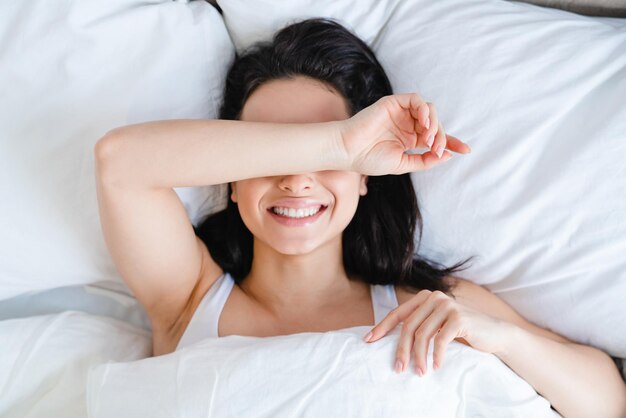 Happy smiling caucasian young woman waking up in her white bed\
in the morning