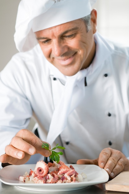 Lo chef sorridente felice guarnisce un'insalata di polpo con una foglia di persil al ristorante