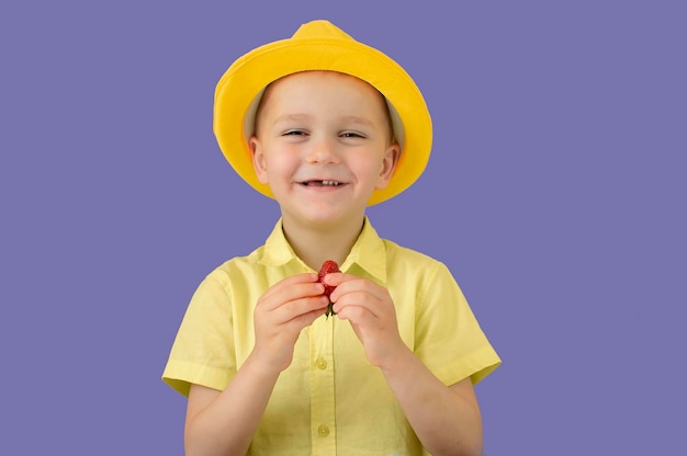 Un bambino sorridente felice con un cappello giallo estivo sta mangiando fragole su uno sfondo viola il concetto di cibo per bambini sano felice infanzia estiva messa a fuoco selettiva