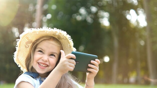 Happy smiling child girl looking in mobile phone outdoors in summer.