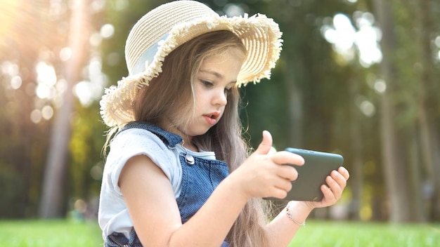 夏に屋外で彼女の携帯電話で見ている幸せな笑顔の子供の女の子。