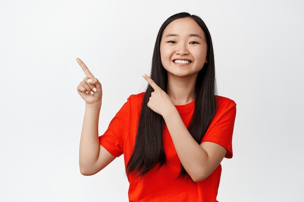 Happy smiling chinese girl pointing fingers at upper left corner, standing in red t-shirt on white