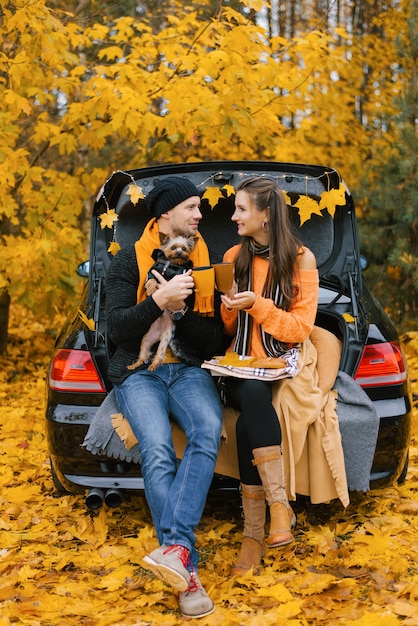 A happy, smiling couple of travelers drink coffee or tea while sitting on the trunk of a car in the autumn forest with their pet