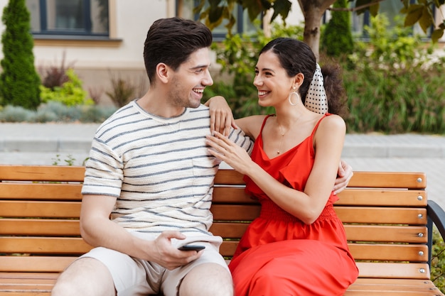 happy smiling couple using cellphone and hugging while sitting on bench in green street