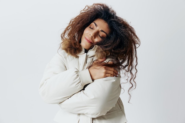 Happy smiling curly pretty latin woman hugging herself enjoyed
beige warm coat closing eyes posing isolated looking at camera at
white studio wall background fashion huge sale new collection
concept